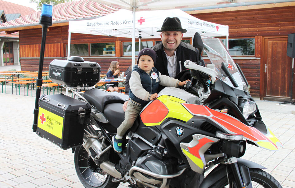 Ein Kind sitzt auf einem Einsatzmotorrad, ein Mann steht dahinter, beide lächeln in die Kamera